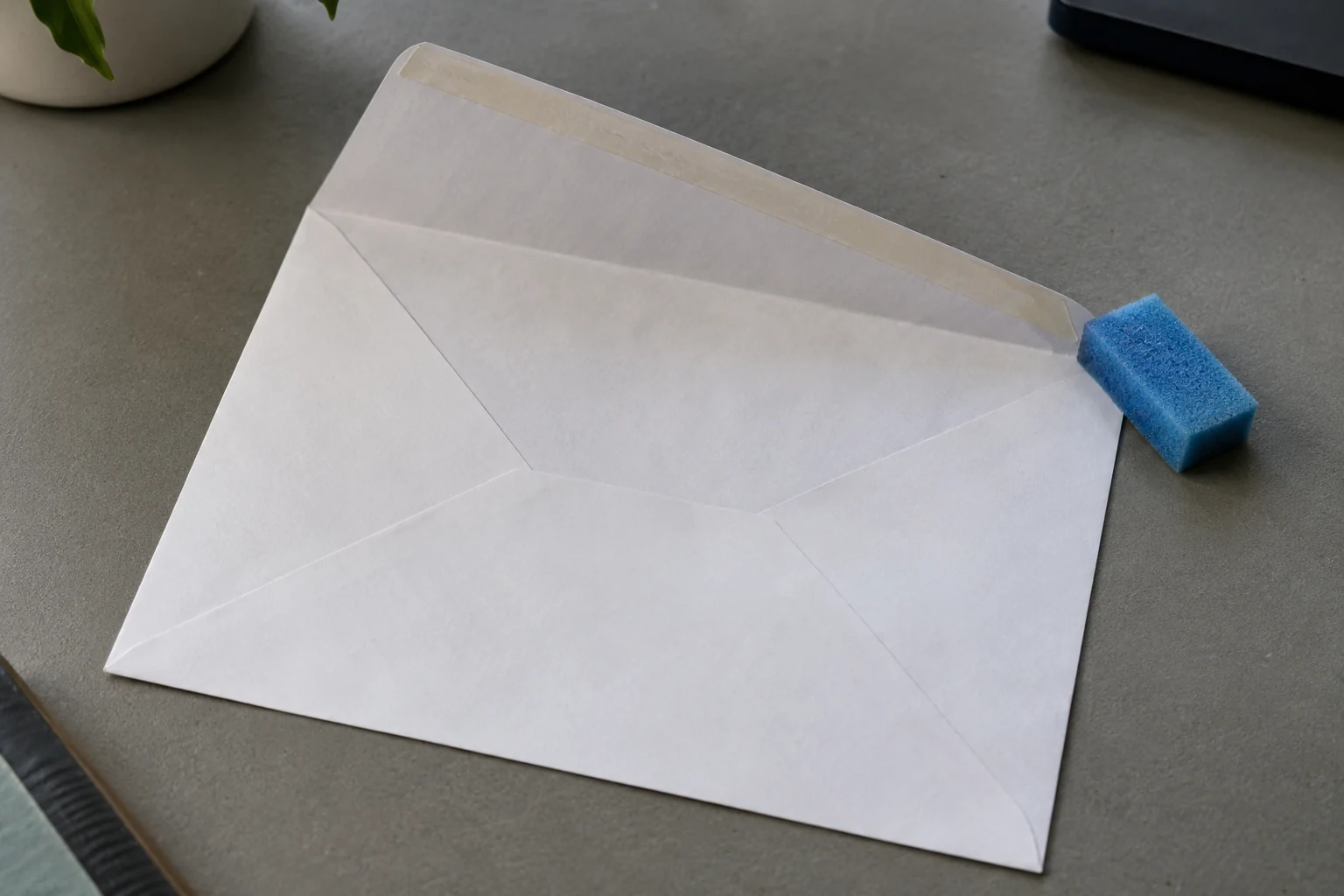 An open white envelope with a pale adhesive band on its flap beside a blue foam moistener on a gray desk.