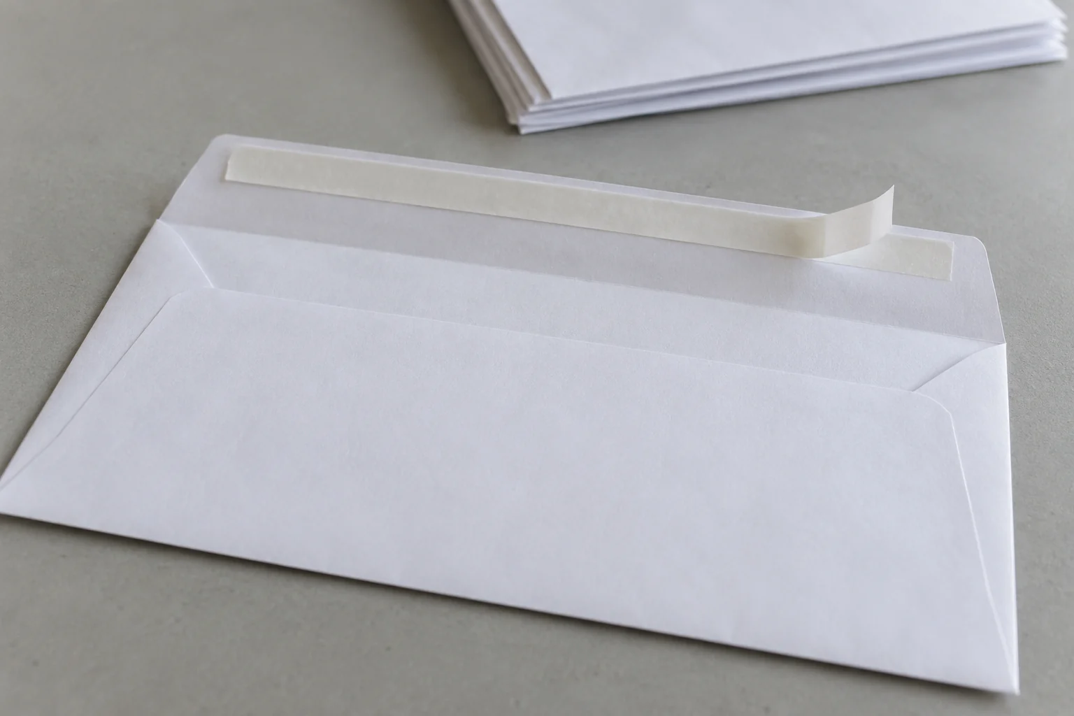 A white envelope with its flap open and a white release strip curled back from one end of the adhesive band, with a small envelope stack behind it.